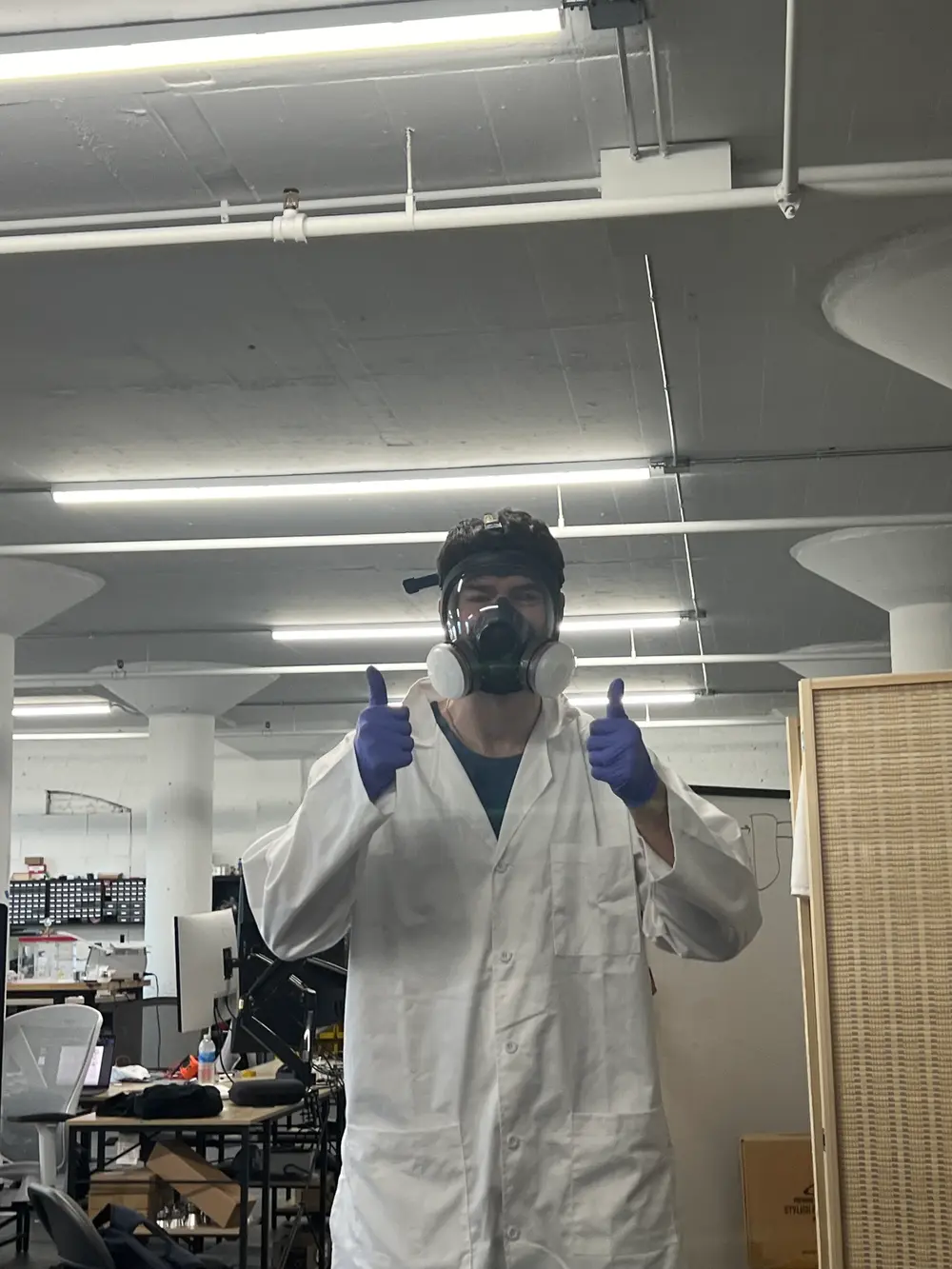 Thumbs up in a lab coat, respirator, and purple nitrile gloves